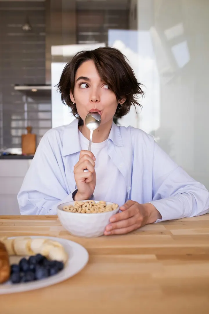 Una chica en su cocina con la cuchara en la boca y un bowl de cereal en la mesa. Fotografía de referencia.